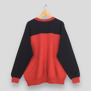 Puede incluir: Una sudadera de dos tonos en negro y rojo cuelga de una percha de madera. La parte superior de la sudadera es negra, con una banda roja horizontal en el pecho. Las mangas son negras y los pu&ntilde;os y el dobladillo inferior son rojos.