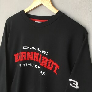May include: Black sweatshirt with the name "DALE EARNHARDT" in white and red lettering. The text "7 TIME CHAMP" is below the name. The number "3" is on the sleeve. The sweatshirt is hanging on a wooden hanger.
