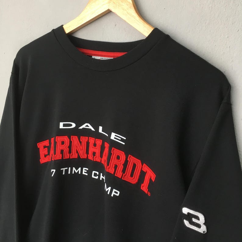 May include: Black sweatshirt with the name "DALE EARNHARDT" in white and red lettering. The text "7 TIME CHAMP" is below the name. The number "3" is on the sleeve. The sweatshirt is hanging on a wooden hanger.