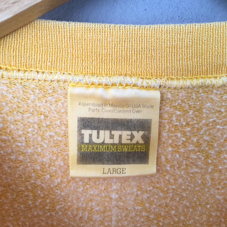 May include: Close-up of a yellow sweatshirt tag. The tag reads "TULTEX MAXIMUM SWEATS LARGE". The tag also states "Assembled in Mexico Or USA Made Parts Care/Content Over". The sweatshirt has a textured, speckled pattern.