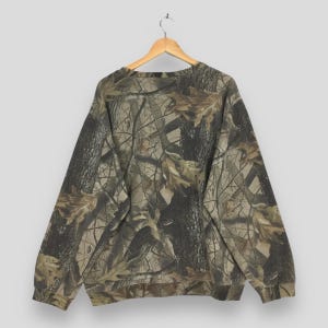 Op de afbeelding: Een sweatshirt met lange mouwen en een ronde hals, voorzien van een realistisch boscamouflagepatroon met donkerbruine boomschorsstructuren en lichtbruine eikenbladeren over de gehele stof. Het kledingstuk hangt aan een houten kledinghanger.