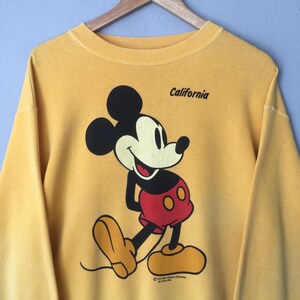 May include: Mustard yellow sweatshirt featuring a smiling Mickey Mouse graphic. The cartoon character is depicted in black, red, and yellow. The word "California" is printed above the graphic. The Walt Disney Company logo is at the bottom.