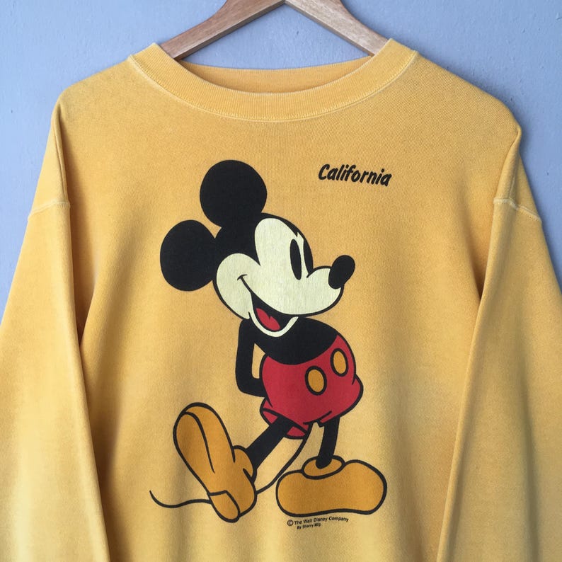 May include: Mustard yellow sweatshirt featuring a smiling Mickey Mouse graphic. The cartoon character is depicted in black, red, and yellow. The word "California" is printed above the graphic. The Walt Disney Company logo is at the bottom.