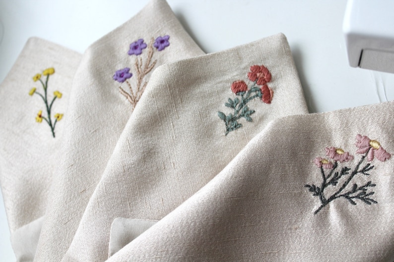 May include: Four linen napkins with embroidered floral designs. Each napkin features a different flower design in various colors, including purple, yellow, orange, and pink.