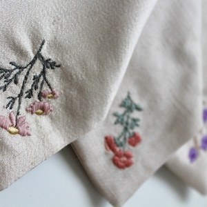 May include: Four fabric squares with embroidered floral designs. The squares are beige, white, and light gray. The flowers are pink, red, green, and purple.
