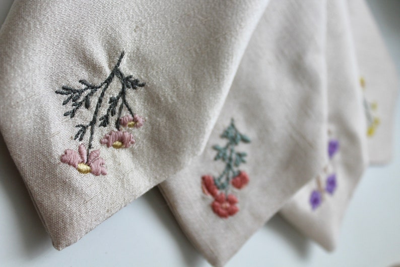 May include: Four fabric squares with embroidered floral designs. The squares are beige, white, and light gray. The flowers are pink, red, green, and purple.