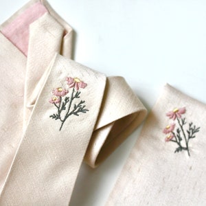 May include: A beige silk tie with a pink lining and a floral embroidery design. The embroidery features pink flowers with yellow centers and gray stems and leaves. A matching pocket square with the same floral embroidery is also shown.
