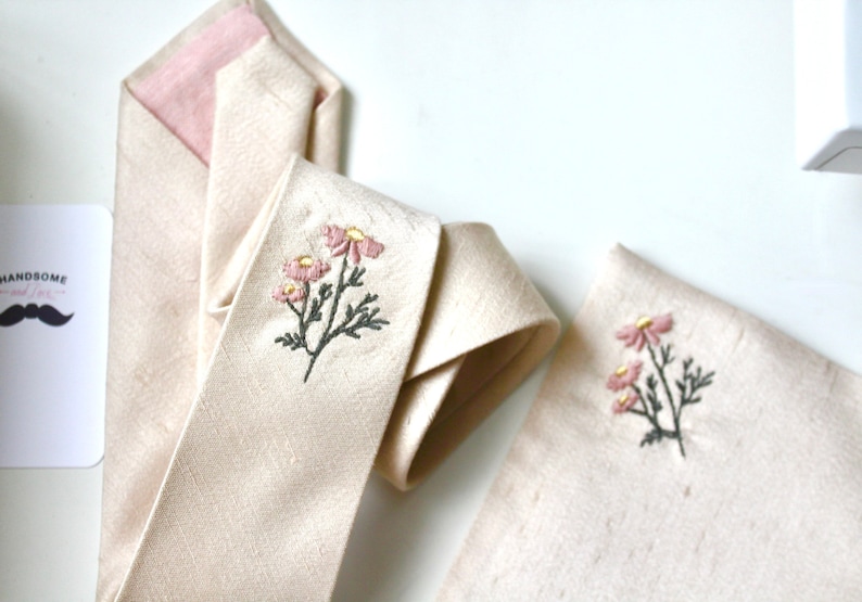 May include: A beige silk tie with a pink lining and a floral embroidery design. The embroidery features pink flowers with yellow centers and gray stems and leaves. A matching pocket square with the same floral embroidery is also shown.