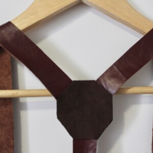 Puede incluir: Un par de tirantes de cuero marr&oacute;n con una pieza trasera en forma de X de cuero marr&oacute;n oscuro. Los tirantes est&aacute;n colgados de una percha de madera.