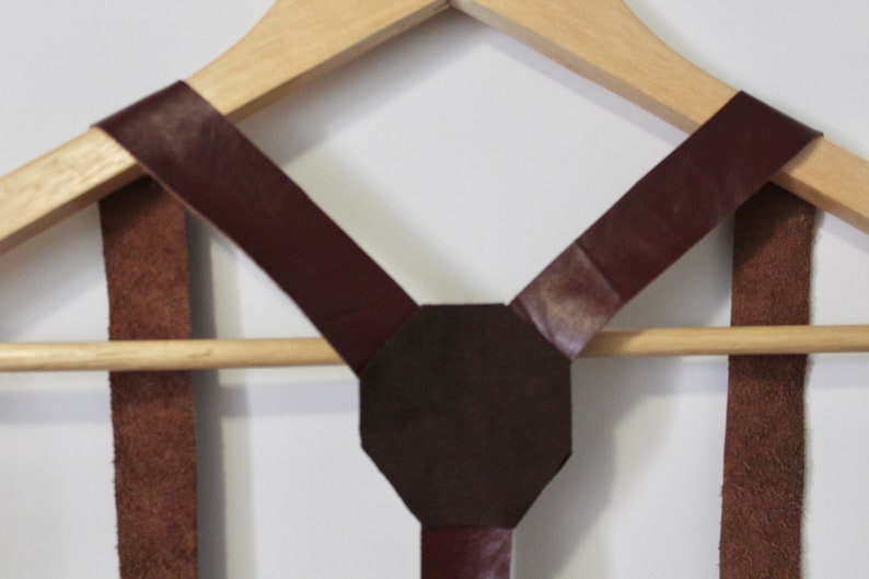 Puede incluir: Un par de tirantes de cuero marr&oacute;n con una pieza trasera en forma de X de cuero marr&oacute;n oscuro. Los tirantes est&aacute;n colgados de una percha de madera.