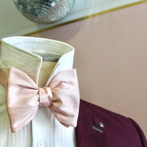 May include: A white dress shirt with a pink bow tie. The shirt has a classic collar and a button-up front. The bow tie is made of a soft, silky fabric.