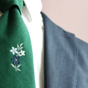 May include: A green tie with a floral embroidery design. The tie is made of a textured fabric and has a knot at the top.