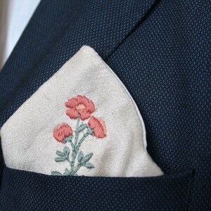 May include: A white linen pocket square with an embroidered floral design featuring pink flowers and green leaves.