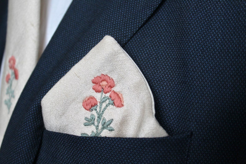 May include: A white linen pocket square with an embroidered floral design featuring pink flowers and green leaves.