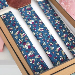 Puede incluir: Un juego de tirantes con un patr&oacute;n floral azul y blanco. Los tirantes est&aacute;n en una caja marr&oacute;n con un parche de cuero.