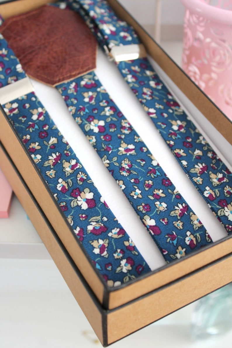 Puede incluir: Un juego de tirantes con un patr&oacute;n floral azul y blanco. Los tirantes est&aacute;n en una caja marr&oacute;n con un parche de cuero.