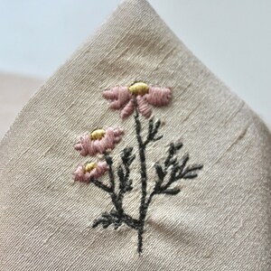 May include: A close-up of a white fabric with a delicate embroidered design of three pink flowers with yellow centers and black stems and leaves.