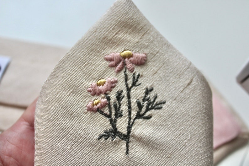May include: A close-up of a white fabric with a delicate embroidered design of three pink flowers with yellow centers and black stems and leaves.