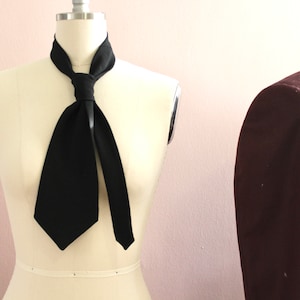 May include: A black necktie tied in a simple knot around a white mannequin's neck. The tie is made of a smooth, silky fabric.