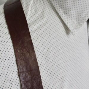 Puede incluir: Primer plano de una tira de tirantes de cuero marr&oacute;n sobre una camisa blanca con un peque&ntilde;o patr&oacute;n de lunares azules.