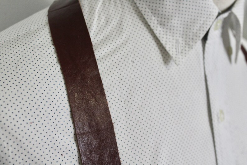 Puede incluir: Primer plano de una tira de tirantes de cuero marr&oacute;n sobre una camisa blanca con un peque&ntilde;o patr&oacute;n de lunares azules.