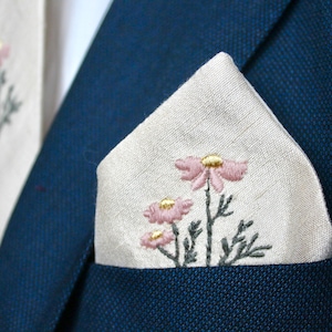 May include: A white linen pocket square with pink and gold embroidered flowers. The pocket square is folded in a triangle shape and placed in the pocket of a navy blue suit jacket.