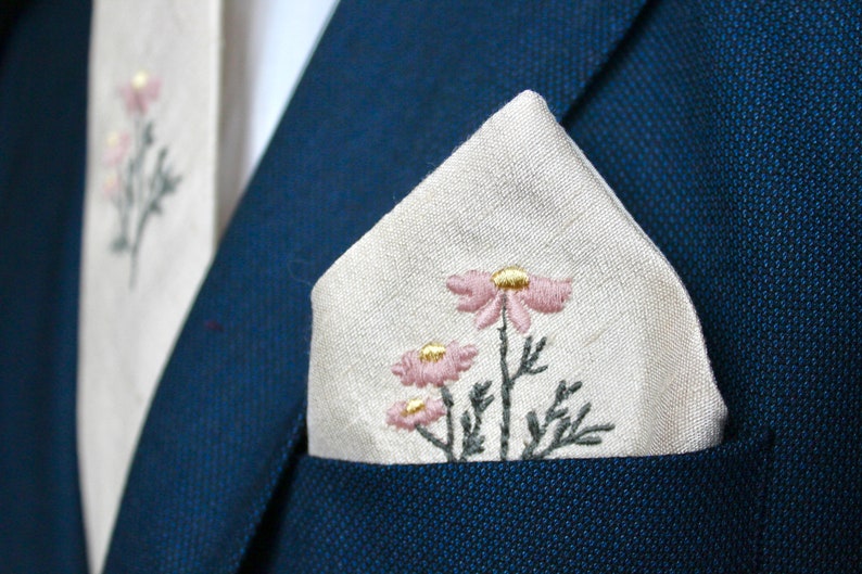 May include: A white linen pocket square with pink and gold embroidered flowers. The pocket square is folded in a triangle shape and placed in the pocket of a navy blue suit jacket.