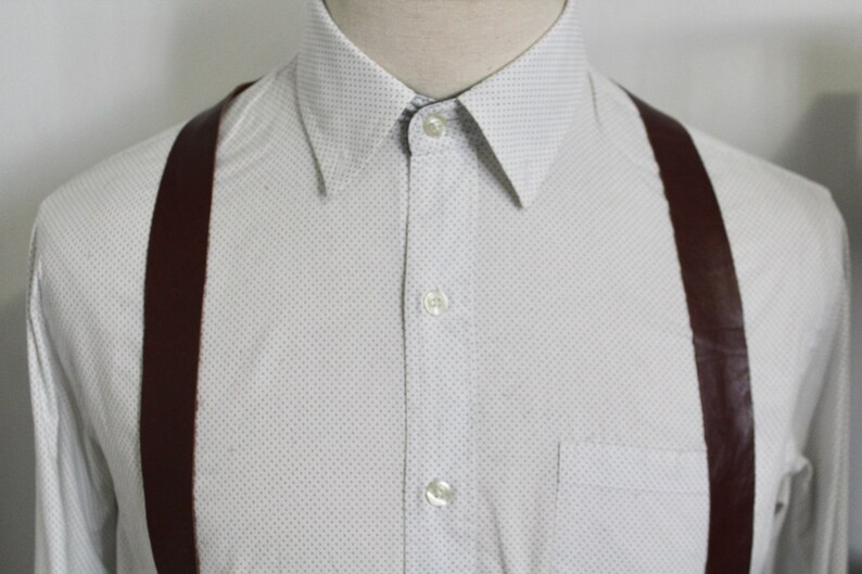 Puede incluir: Una camisa blanca de botones con un peque&ntilde;o patr&oacute;n de puntos blancos y tirantes marrones.