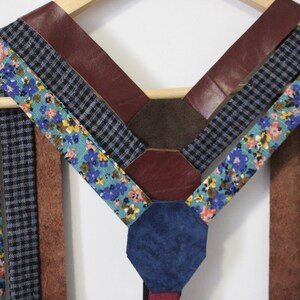 Puede incluir: Un conjunto de tirantes hechos de diferentes telas, incluyendo tela con estampado floral, tela a cuadros y cuero marr&oacute;n. Los tirantes est&aacute;n dispuestos en forma de V y est&aacute;n colgados de una percha de madera.