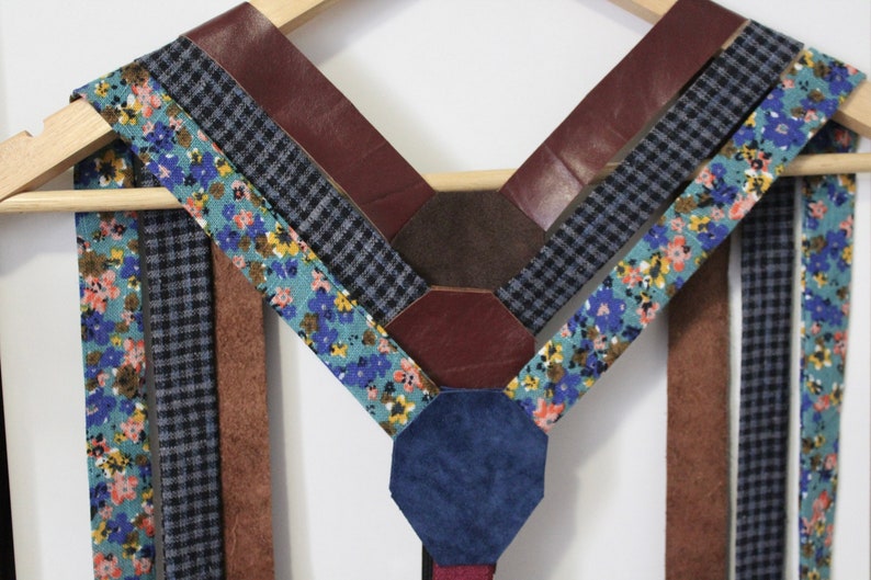 Puede incluir: Un conjunto de tirantes hechos de diferentes telas, incluyendo tela con estampado floral, tela a cuadros y cuero marr&oacute;n. Los tirantes est&aacute;n dispuestos en forma de V y est&aacute;n colgados de una percha de madera.