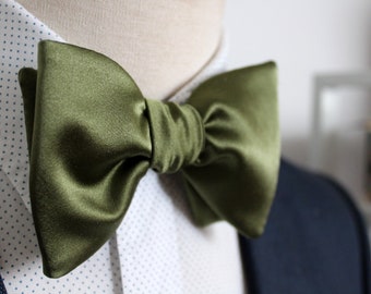 Puede incluir: Pajarita de satén verde oliva. La pajarita está atada y colocada sobre una camisa blanca con un estampado de lunares azules. La pajarita es el punto focal, con un brillo suave y lustroso.