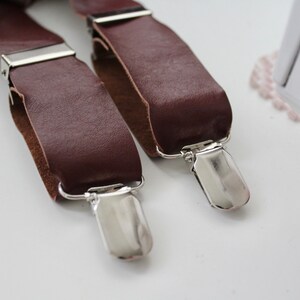 Puede incluir: Dos tirantes de cuero marr&oacute;n con clips de metal plateado. Los tirantes est&aacute;n sobre una superficie blanca.