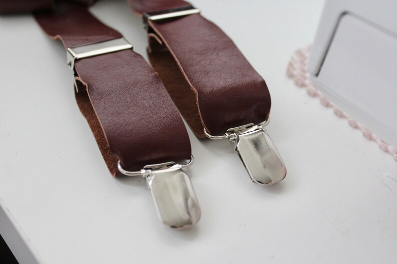Puede incluir: Dos tirantes de cuero marr&oacute;n con clips de metal plateado. Los tirantes est&aacute;n sobre una superficie blanca.