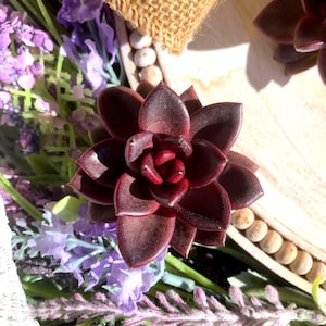 May include: A close-up of a dark burgundy succulent plant with layered petals. The succulent is surrounded by purple lavender flowers and a wooden tray with white beads. The lighting creates shadows, highlighting the plant's texture and color.