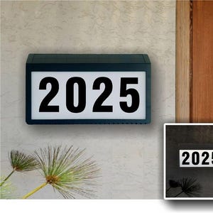 May include: A black rectangular house number sign with a white background and the number 2025 displayed in black. The sign is mounted on a light gray wall.