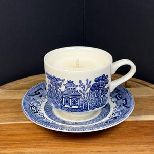 May include: A white teacup and saucer set with a blue Willow pattern, filled with a cream-colored candle. The candle has a single wick. The teacup sits on the saucer, both on a wooden surface.