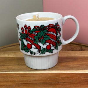 May include: A white ceramic mug candle with a Christmas bell design. The mug features red bells, green holly, and red ribbons. The candle has a beige wax and a single wick. The mug sits on a white, ribbed base.