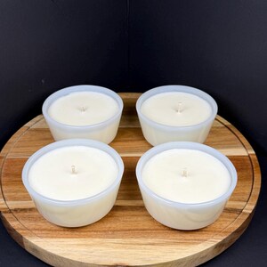 May include: Four white candles in small, round, white containers, each with a single wick. The candles sit on a round, wooden serving board with a light brown finish. The background is a solid black.