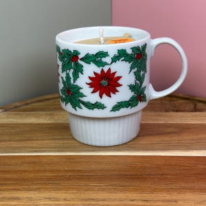 May include: A white ceramic mug candle with a festive Christmas design. The mug features a red poinsettia flower and green holly leaves. The candle has a white wick and is set on a wooden surface.