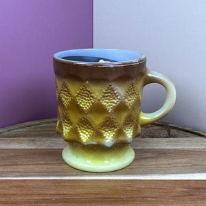 May include: A yellow and brown ceramic mug with a textured diamond pattern. The mug has a light green base and handle, with a dark brown rim. The mug is sitting on a wooden tray.