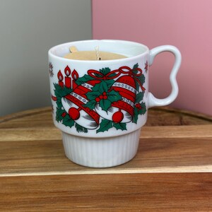 May include: White ceramic mug with a Christmas bell design in red and green. The mug has a handle and a ridged base. The mug is filled with a candle. The candle has a wooden wick and a brown paper label.