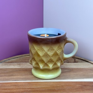 May include: A vintage-style candle in a mug. The mug is a light yellow-green color with a diamond pattern and a brown gradient. The candle wax is black, and the interior of the mug is light blue.