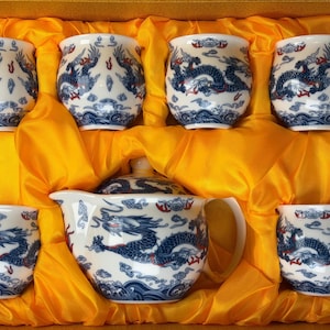 May include: A white porcelain tea set with a blue dragon design, including a teapot, a lid, and six cups. The set is nestled in a yellow satin-lined box, creating a visually appealing presentation.