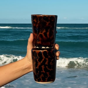 Può includere: Due tazze cilindriche in ceramica impilate verticalmente, caratterizzate da una smaltatura marrone lucida con un motivo leopardato nero scuro. Le tazze presentano un bordo arrotondato liscio e una base chiara visibile sul fondo.
