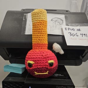 May include: A crocheted red, orange, and yellow bong with a white crocheted lid and black eyes. The bong is sitting on a black printer with a blue sticky note on the side. The text "EPOD # 306 491 (Back Hallway" is written on a white sticky note.
