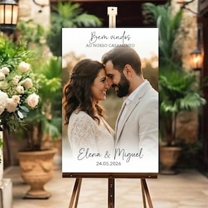 Op de afbeelding: Een rechthoekig welkomstbord voor een bruiloft op een metalen ezel met een foto van een koppel. De tekst luidt Bem vindos AO NOSSO CASAMENTO, Elena & Miguel en 24.05.2026. Het bord heeft een witte achtergrond met zwarte schreefletters.