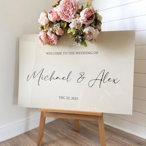 May include: A wedding welcome sign with the text "WELCOME TO THE WEDDING OF Michael & Alex DEC 22, 2025" in elegant script. The sign is cream-colored and displayed on a wooden easel, adorned with pink and white floral arrangement.