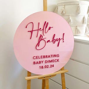 Może przedstawiać: Okrągła tabliczka z jasnoróżowego akrylu z ciemnoróżowymi wypukłymi literami o treści Hello Baby! CELEBRATING BABY DIMECK 18.02.24. Tabliczka jest umieszczona na jasnej drewnianej sztaludze, prezentując tekst w połączeniu czcionki pisanej i blokowej.