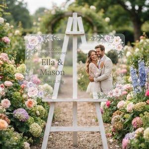 May include: A clear acrylic wedding welcome sign on a white wooden easel, featuring floral illustrations in pink and purple, with the text "WELCOME TO OUR WEDDING", "Ella & Mason", and "October 12, 2024", alongside a photo of a couple.