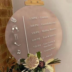 Peut inclure: Panneau de mariage rond, mauve, avec un programme d'événements en texte blanc. Des icônes décoratives sont incluses. Le panneau est soutenu par un chevalet en bois, avec des décorations florales à la base.